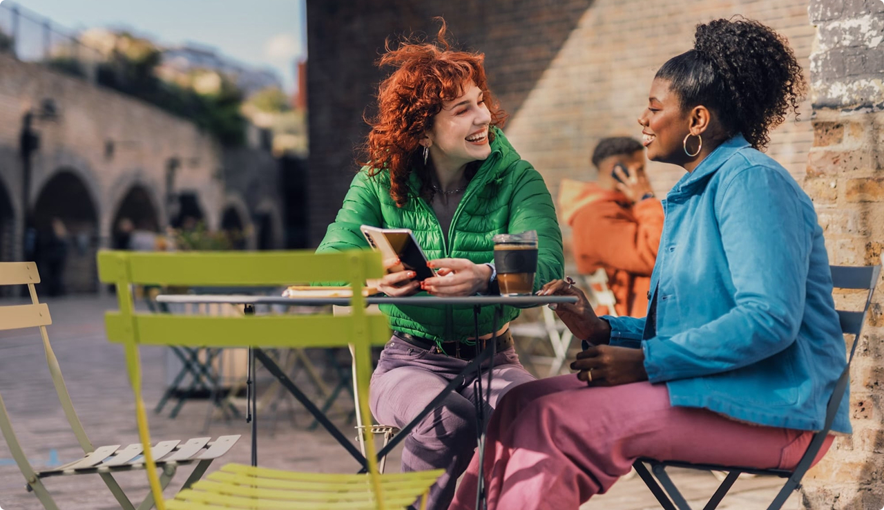 Three women chatting at an outdoor caf&eacute; with colorful chairs and city buildings in background.