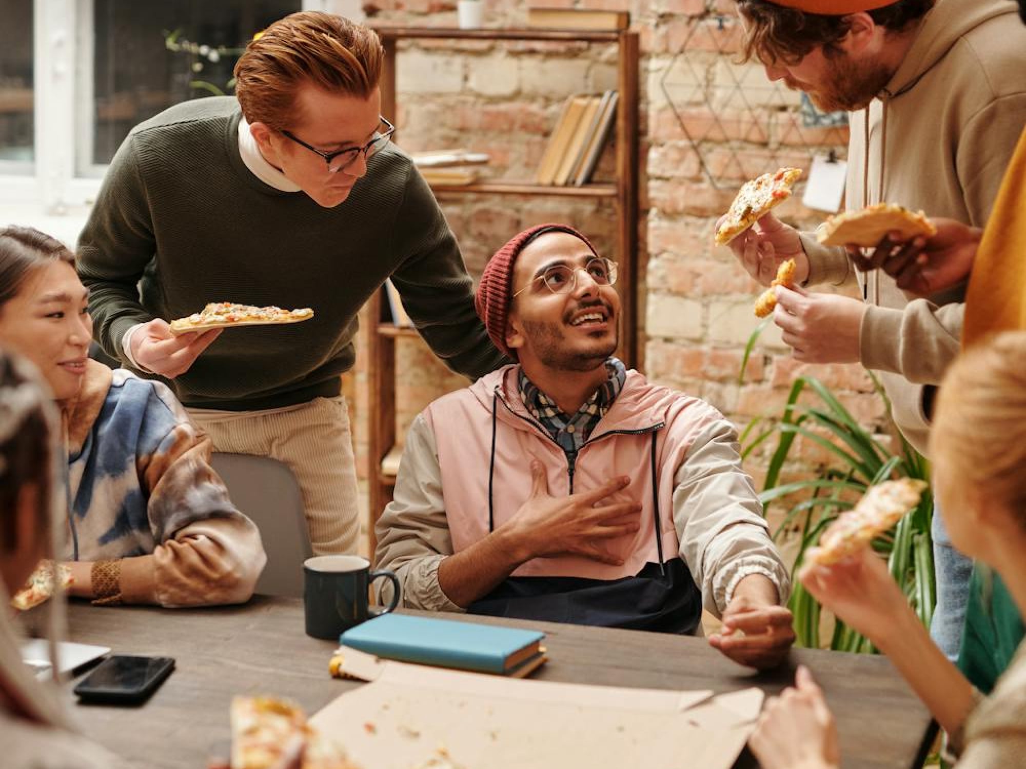 Coworkers laughing over pizza at a meeting table in a warm brick-walled office