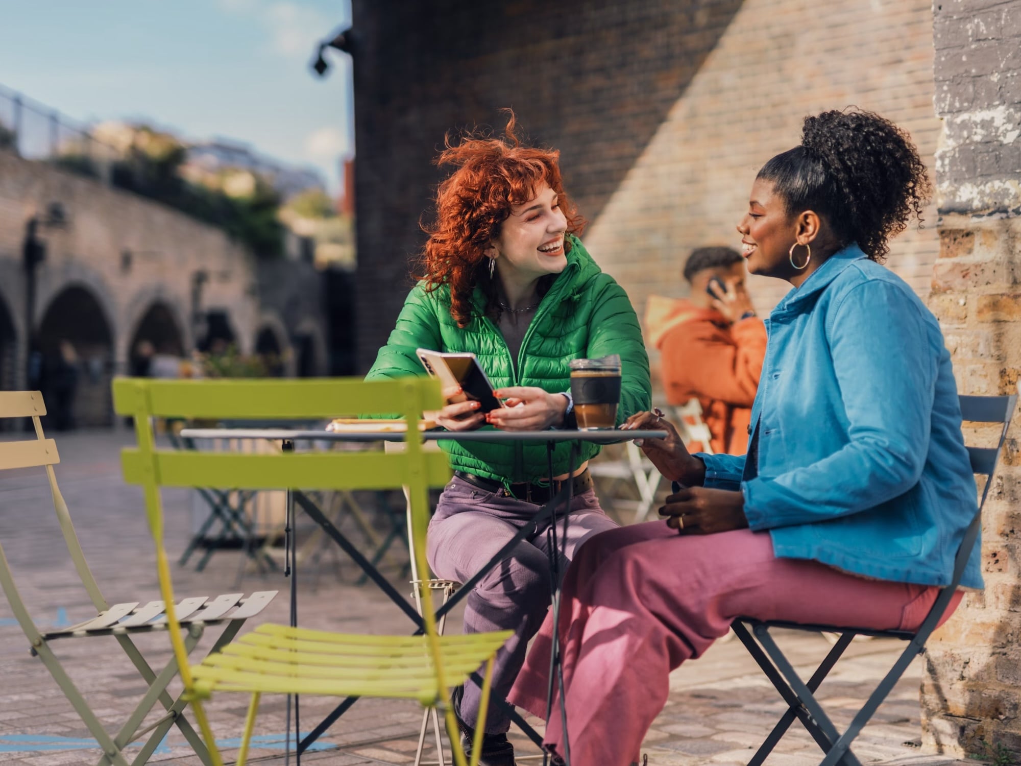 Two women chatting and smiling at an outdoor caf&eacute; table, drinking tea on sunny day